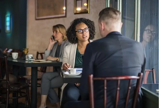 a business man and woman sitting at a table where the woman looks taken aback and there is a woman on the phone in the background