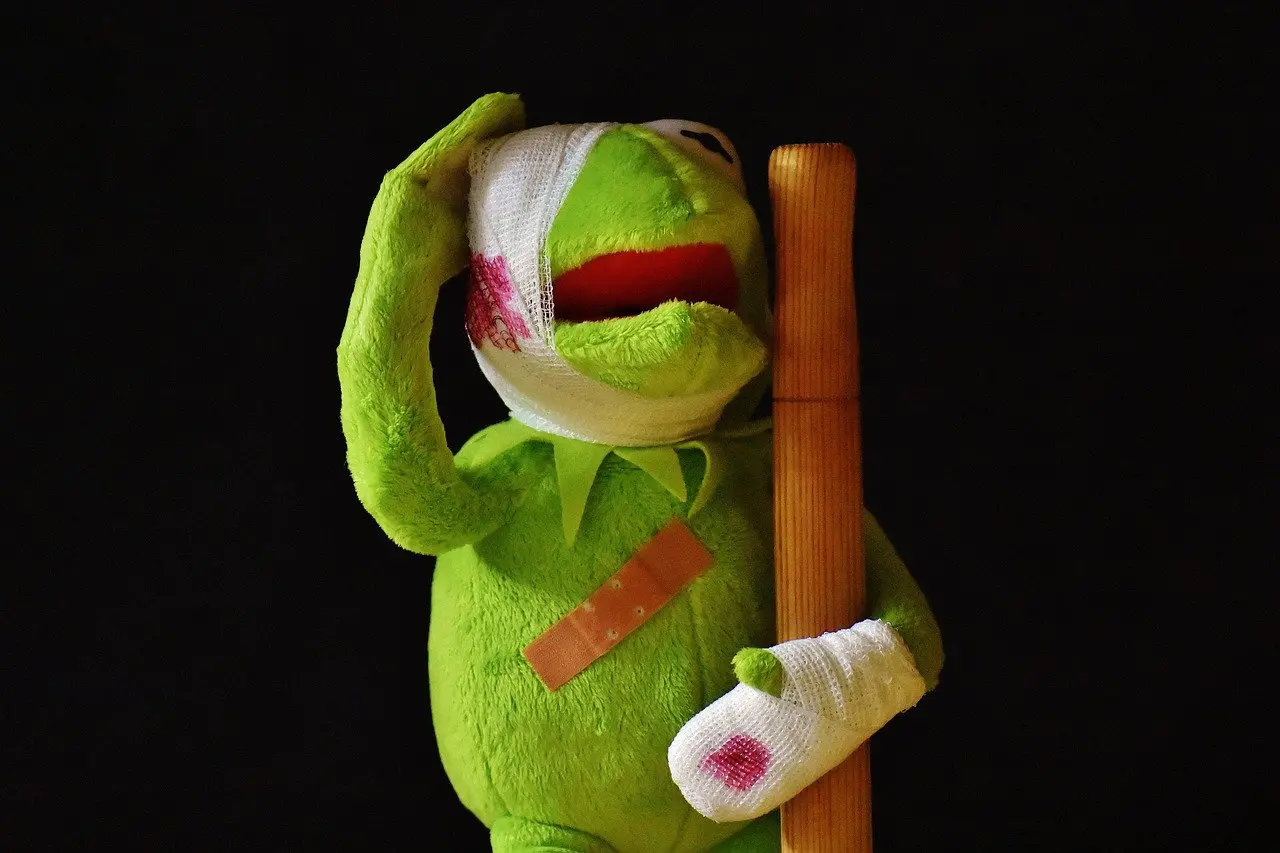 a bruised and bandaged Kermit the frog holding a wooden stick