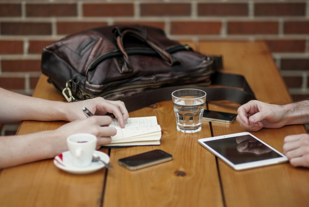 two individuals' hands, sat at a table, one is writing in a notebook, the other had a tablet in front of them