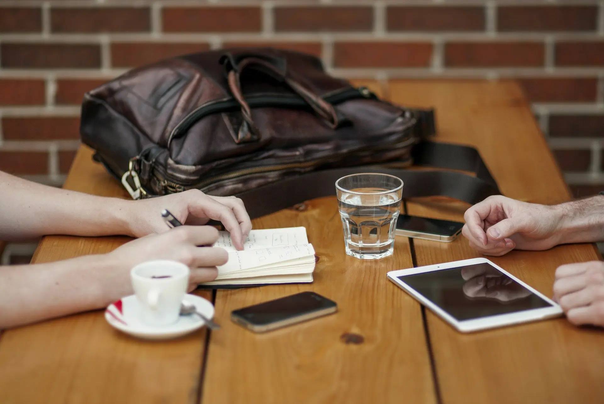 two individuals' hands, sat at a table, one is writing in a notebook, the other had a tablet in front of them