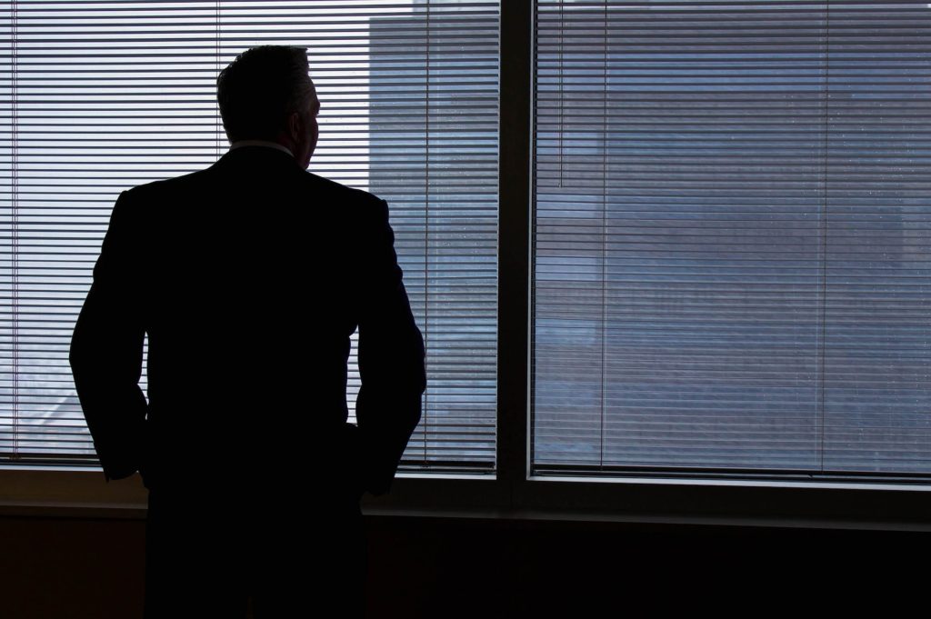 man in a suit stands in front of a window with blinds