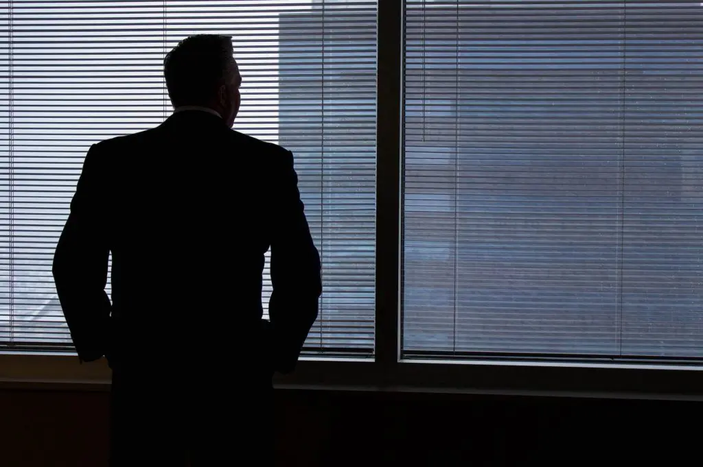 man in a suit stands in front of a window with blinds