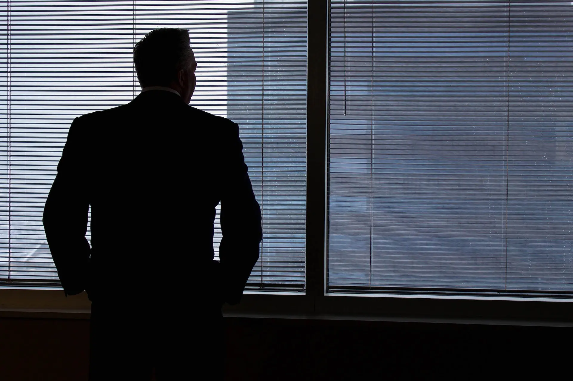 man in a suit stands in front of a window with blinds