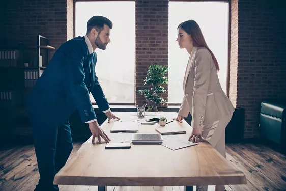 Business man and woman standing at a table looking at each other & discussing disagreement