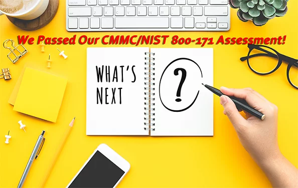 the text 'we passed our CMMC/NIST 800-171 assessment!' and the question 'what's next?' on a notebook with a hand hovering holding a pen