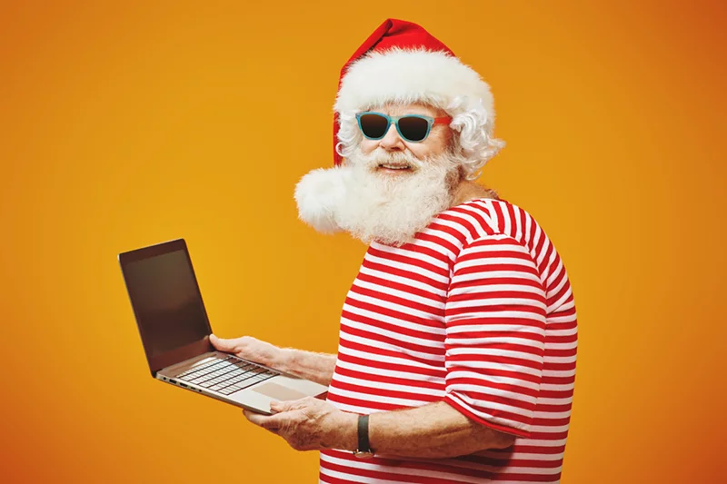 an individual dressed like Santa in a red and white striped shirt with a red hat is holding a laptop